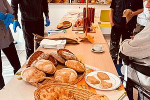Mehrere Personen stehen an einem Tisch mit verschiedenen Brotsorten und Gebäck in einem hellen Raum