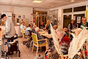 Senioren sitzen an Tischen in einem hellen Gemeinschaftsraum, im Vordergrund ein Tisch mit Muffins und dekorativen Vasen mit Pampasgras
