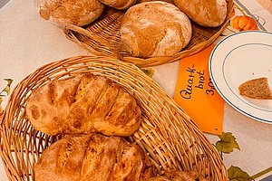 Zwei geflochtene Körbe mit verschiedenen Brotsorten auf einem Tisch, daneben ein Teller mit einem Stück Brot und zwei orangefarbene Zettel mit handgeschriebenen Texten