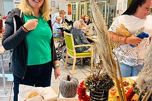 Frau in grünem Shirt und schwarzer Jacke steht lächelnd an einem Tisch mit Muffins und herbstlicher Dekoration in einem Raum mit mehreren sitzenden älteren Menschen.