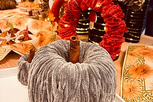 Kuscheliger grauer Kürbis aus Stoff mit braunem Stiel auf einem weißen Tisch vor herbstlicher Dekoration mit roten und orangefarbenen Elementen