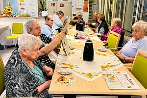 Mehrere ältere Menschen sitzen an einem langen Tisch in einem hellen Raum und lesen Zeitungen oder unterhalten sich.