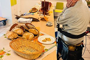 Älterer Mann mit schwarzer Kappe sitzt auf einem Rollator an einem Tisch mit Körben voller Brot und Gebäck in einem hellen Raum